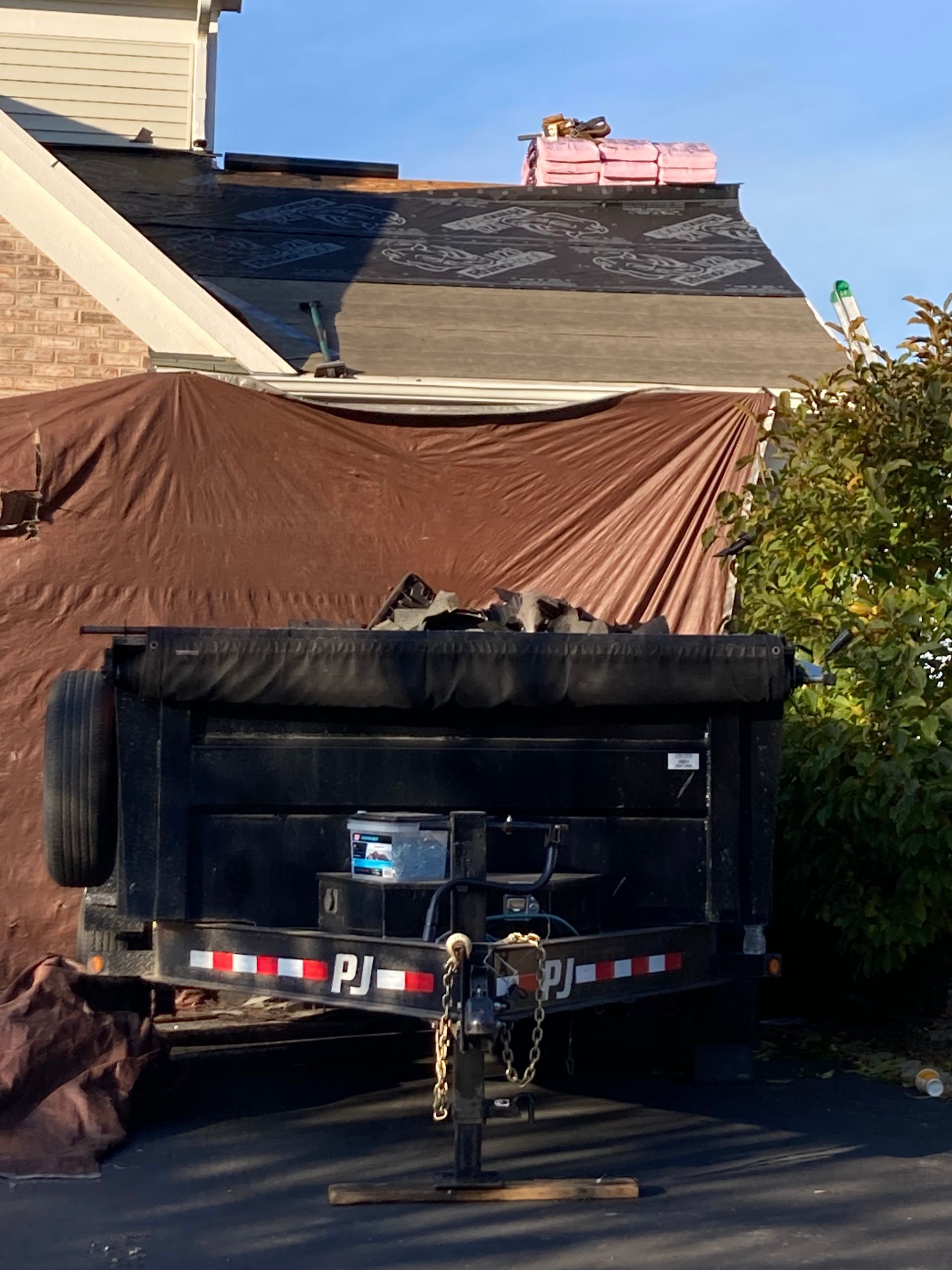 Roof replacement in progress: tarp covers debris, dumpster in foreground, shingles stacked.