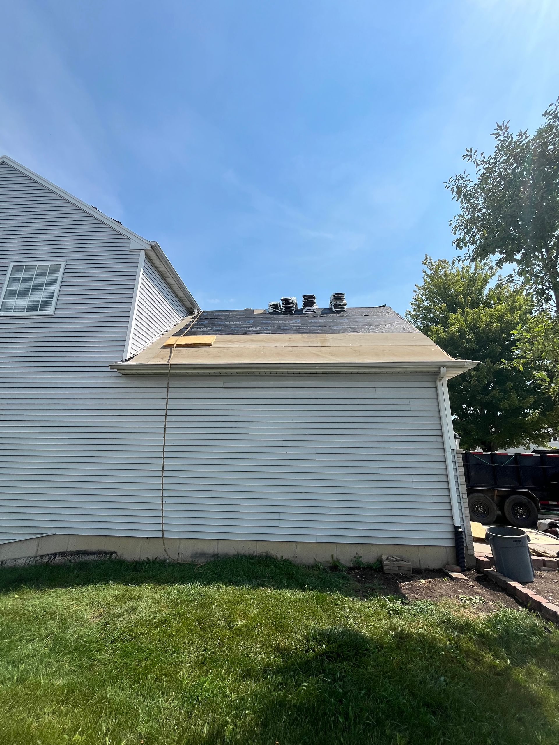 Partially shingled roof and siding on a house under a blue sky, grass lawn in the foreground.