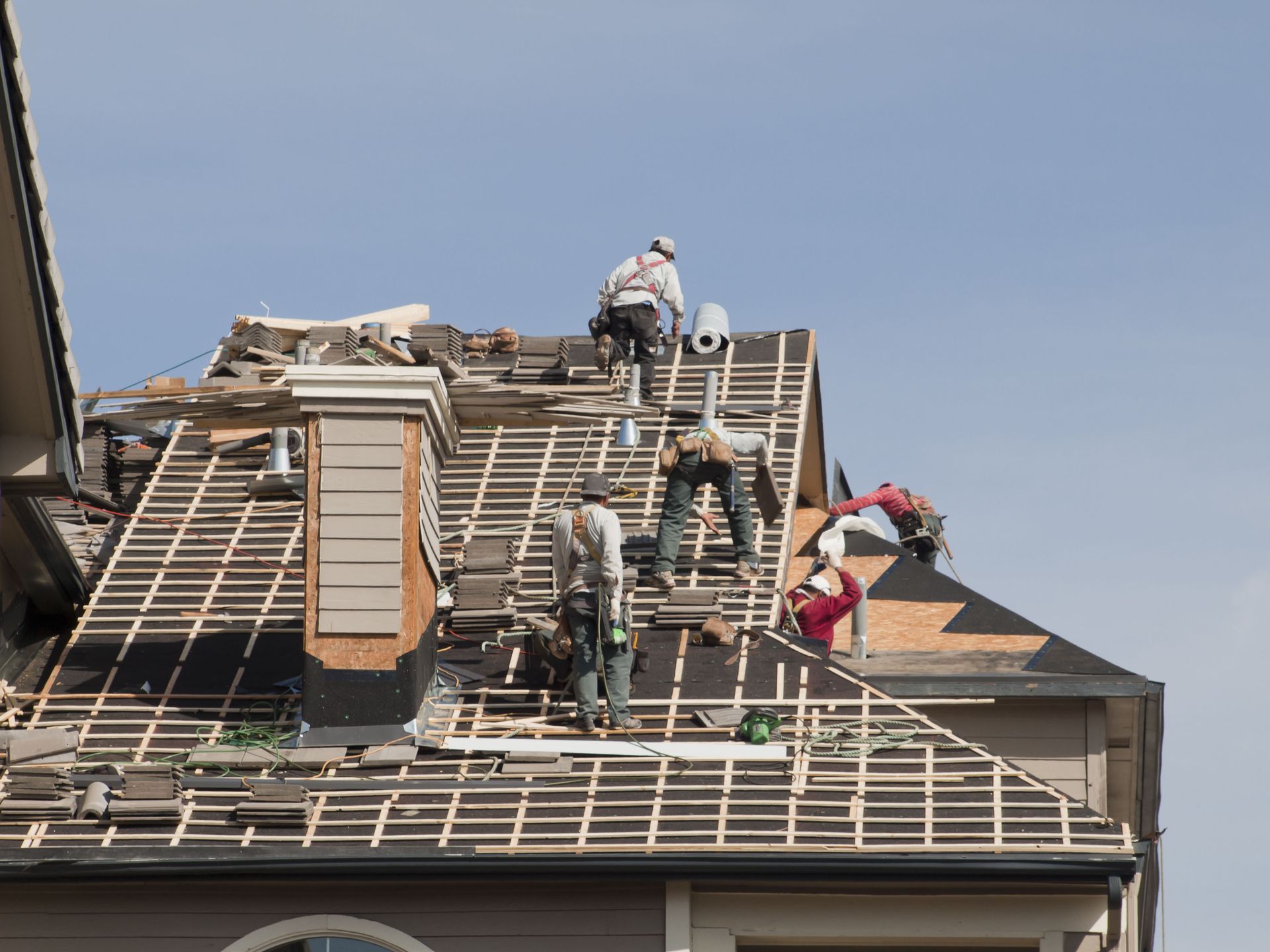 Roofers working on a house roof under a blue sky, removing old shingles and installing new ones.