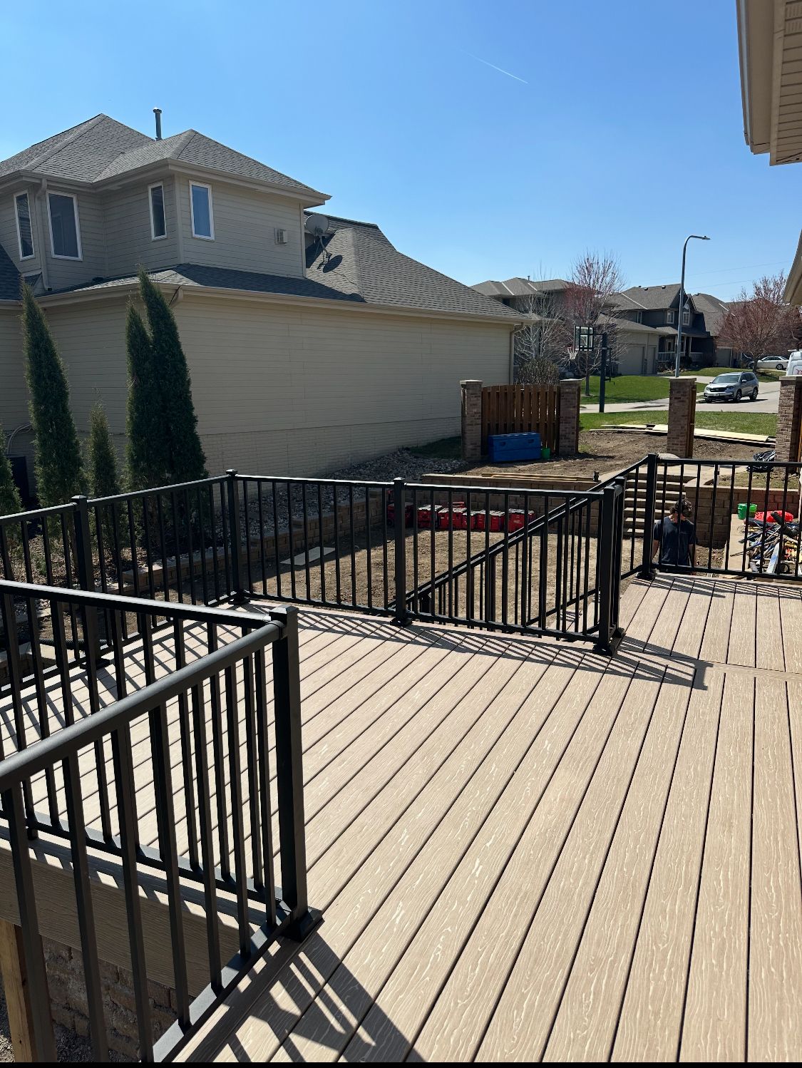 Wooden deck with black railing, overlooking a yard with a house in the background on a sunny day.