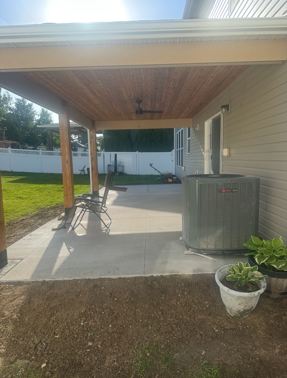 Covered patio with wooden ceiling, concrete floor, next to a house with an AC unit and potted plants.