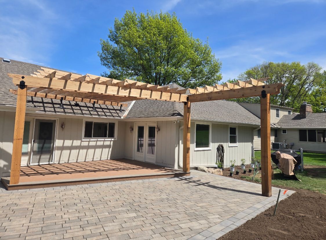 Wooden pergola over a brick patio adjacent to a house. Sunny day with green trees in the background.