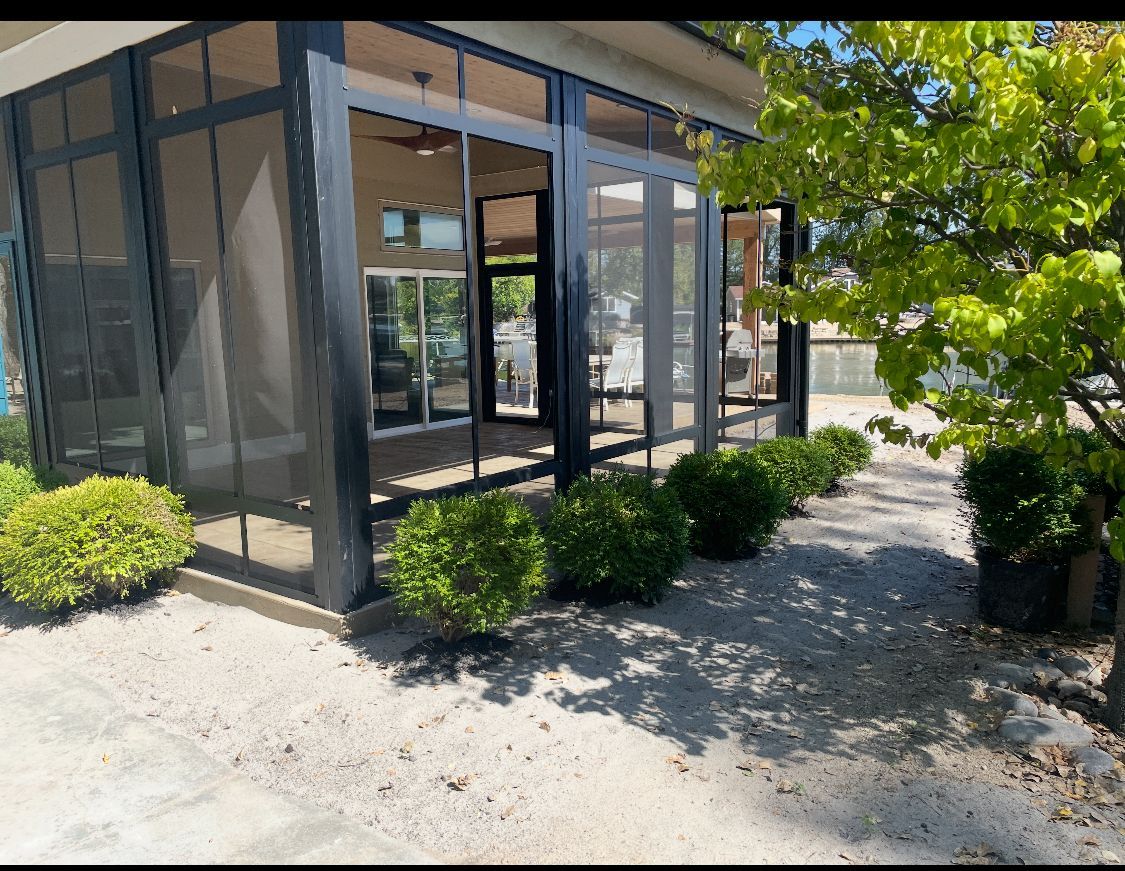 Screened-in patio with black frames and glass panels, surrounded by bushes and a tree. Gravel path.