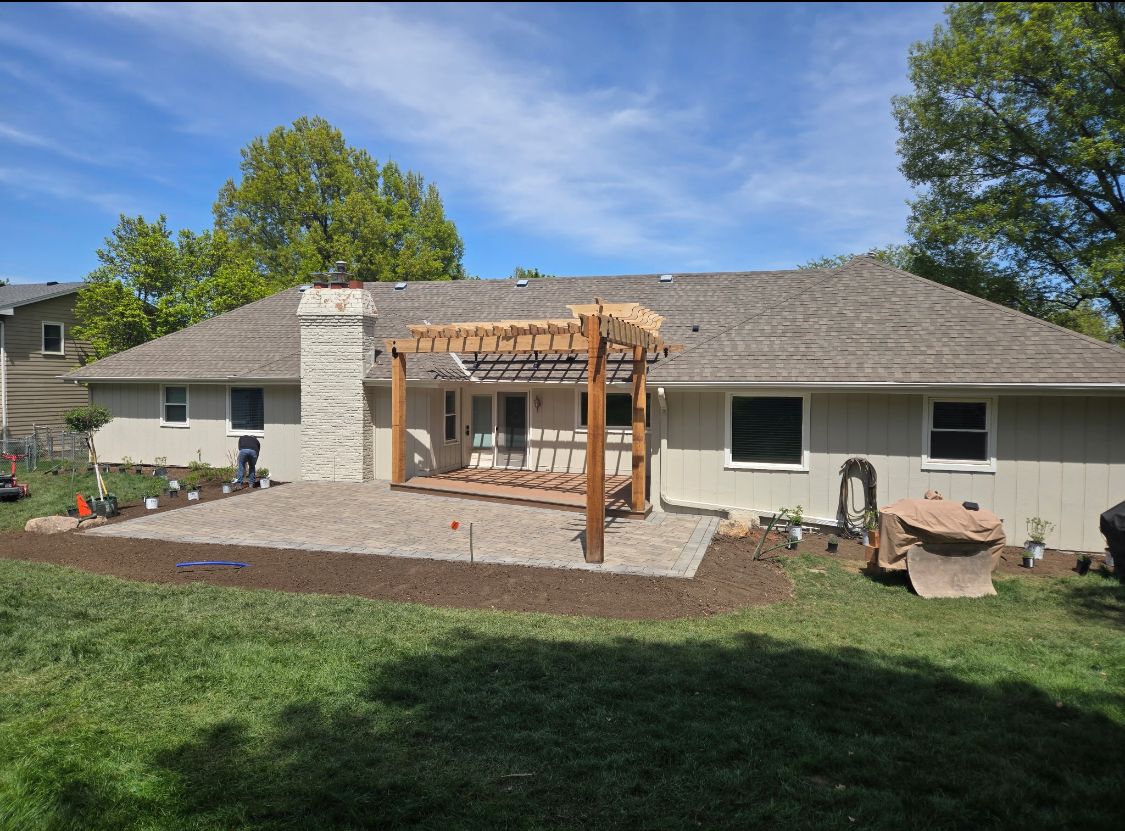 Backyard with a gravel patio, wooden pergola, and house. Green grass and blue sky are in the background.
