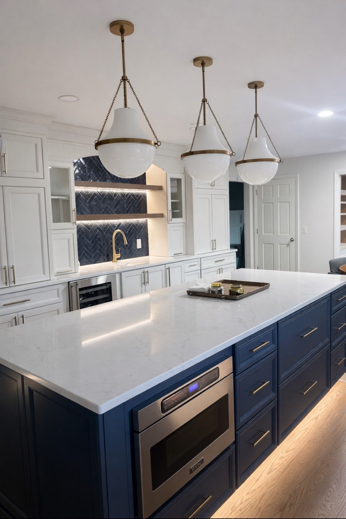 Modern white kitchen with navy island, marble countertop, pendant lights, and built-in oven