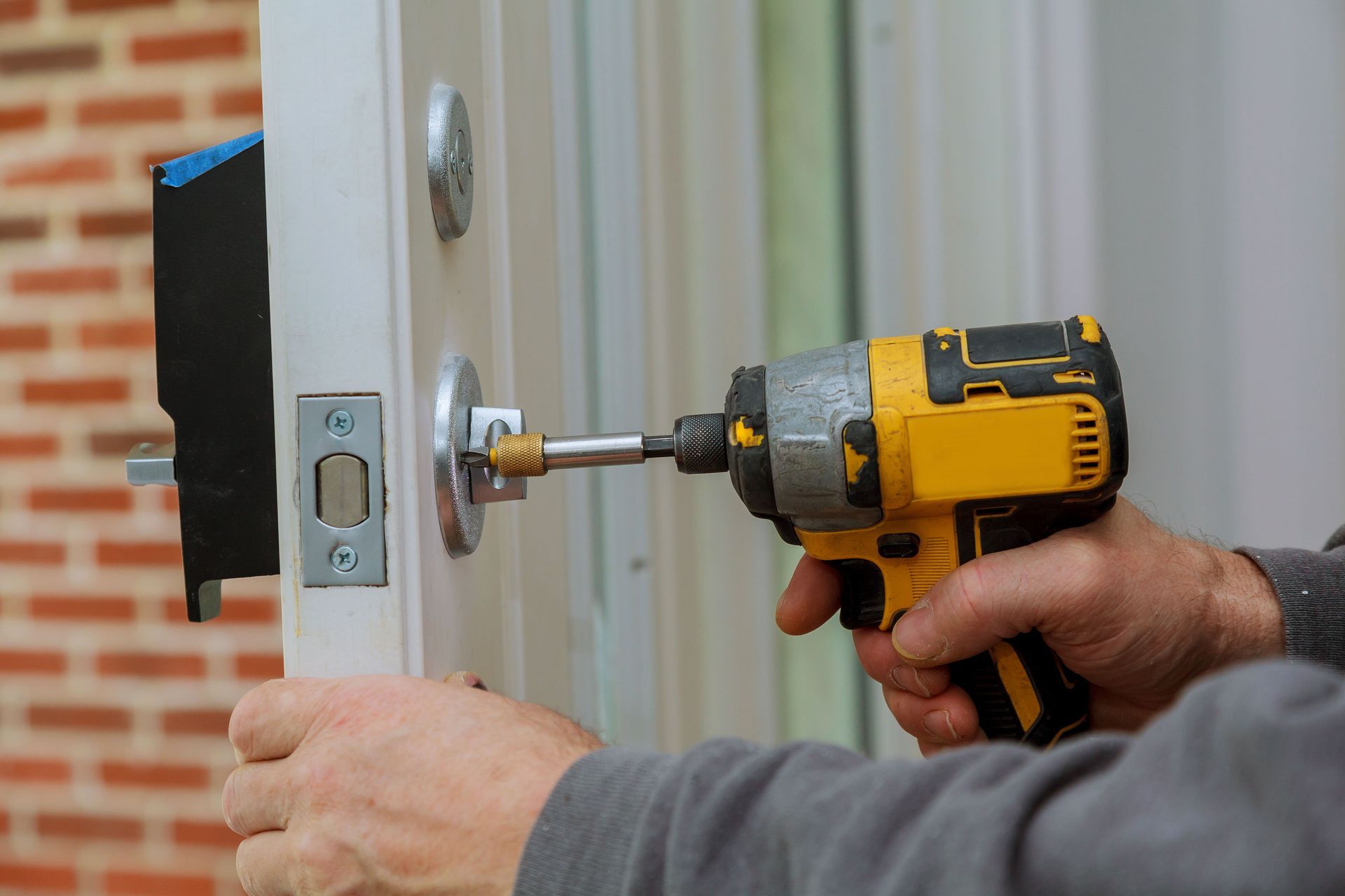 Person using a yellow drill to install a silver door lock on a white door. Brick wall in the background.