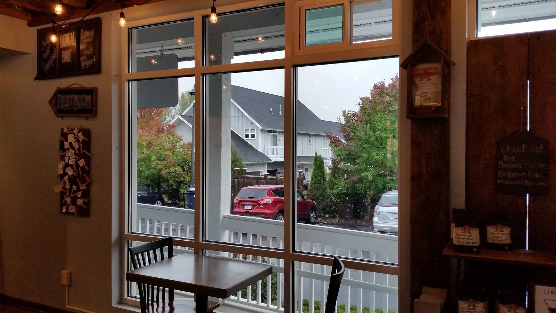 Interior view of a cafe with a window overlooking a street with houses and cars; a table and chairs sit near the window.