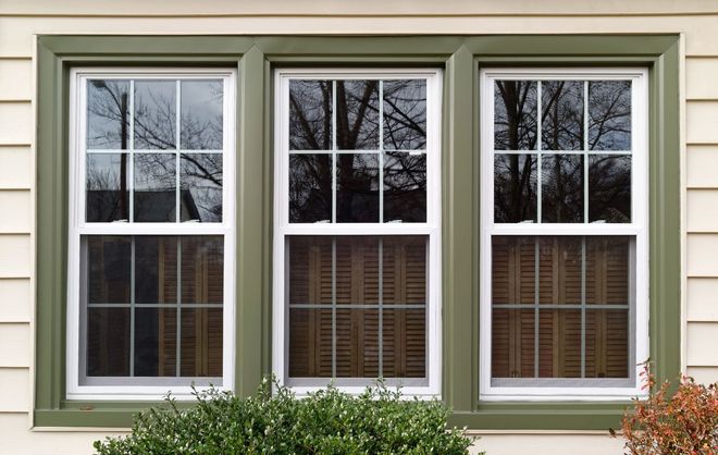 Three white-framed windows with green trim reflect trees and have blinds partially visible. The wall is beige.