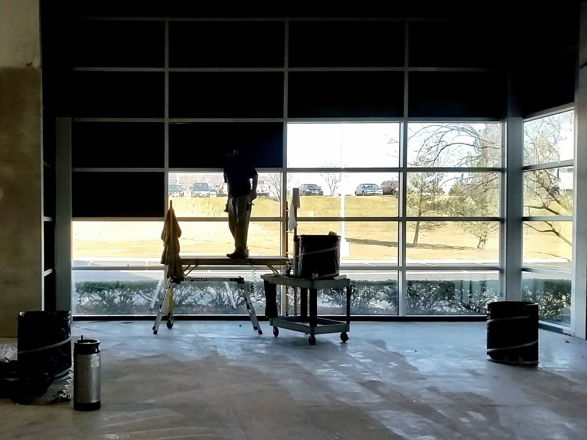 A person on a platform installs a black window shade in a building with large windows overlooking a grassy area.