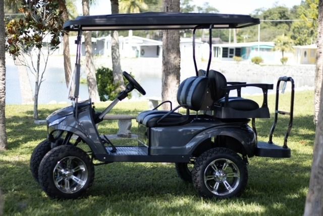 Gray golf cart with lifted wheels, parked on grass near water and trees.