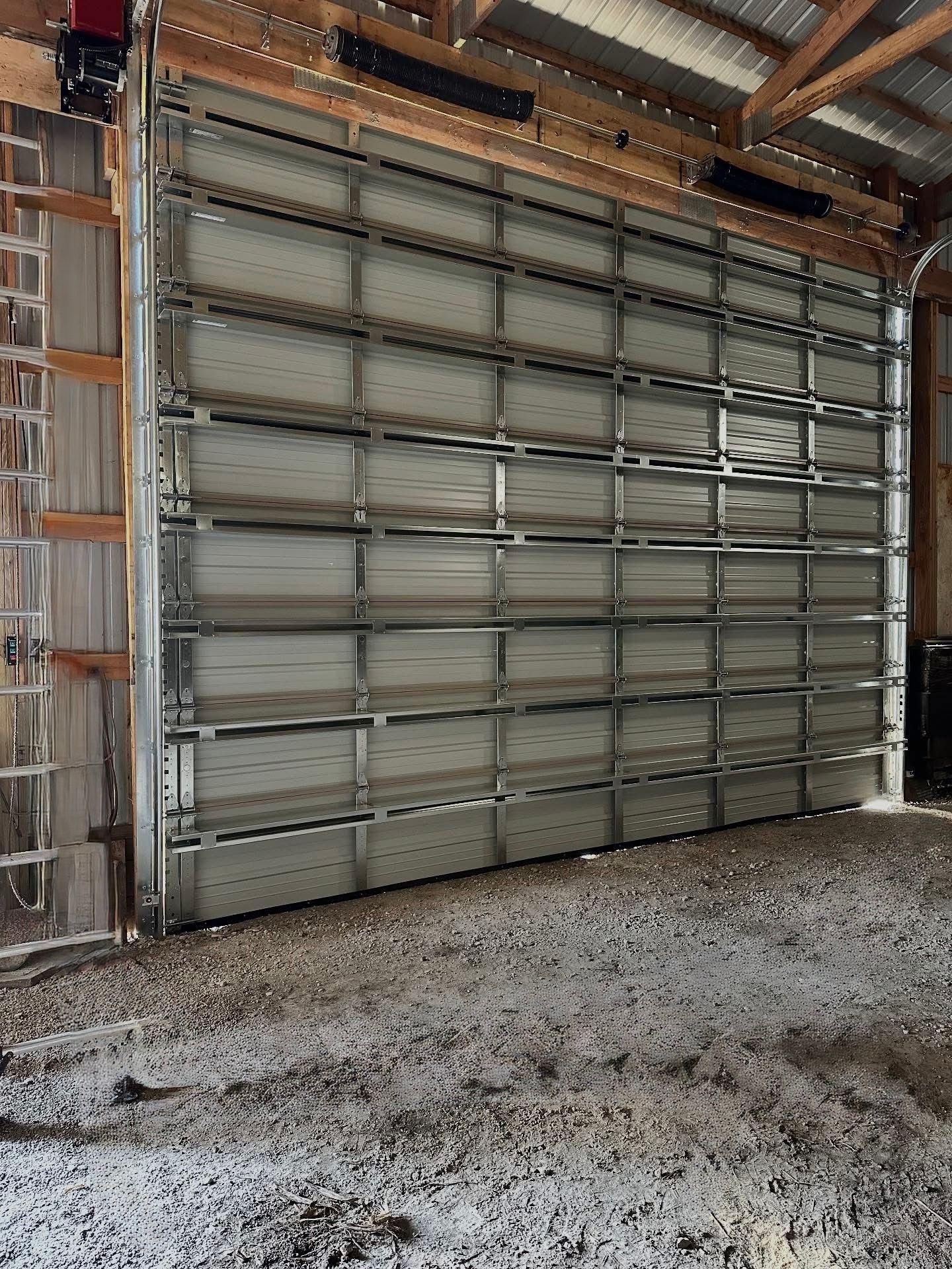 Large metal overhead garage door inside a barn. Door is closed, with a gravel floor below.