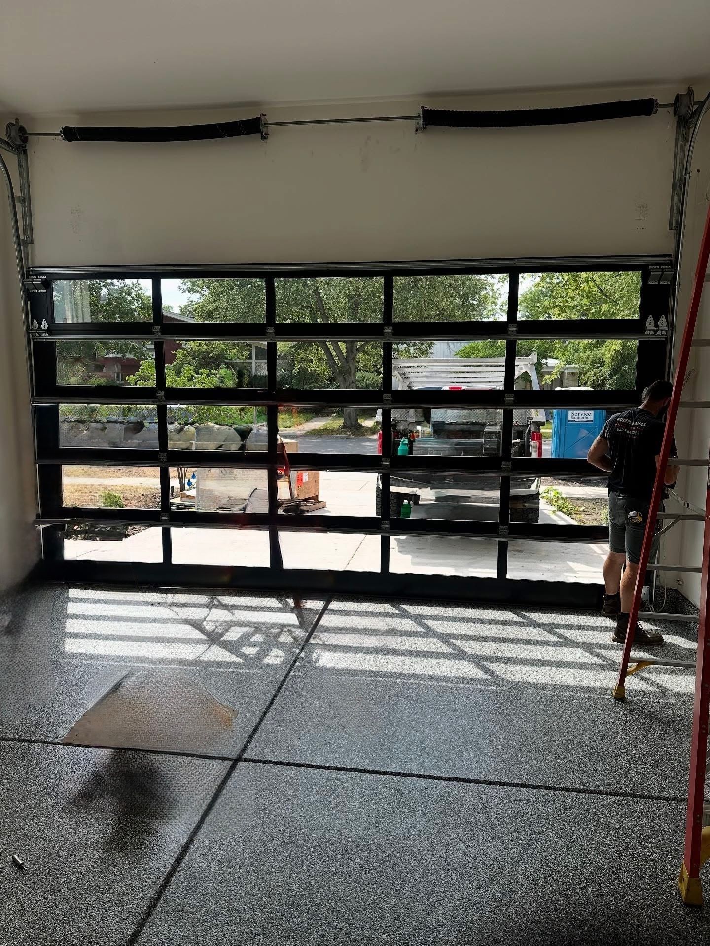 Garage door with black framing and clear panels; a person on a ladder next to it.