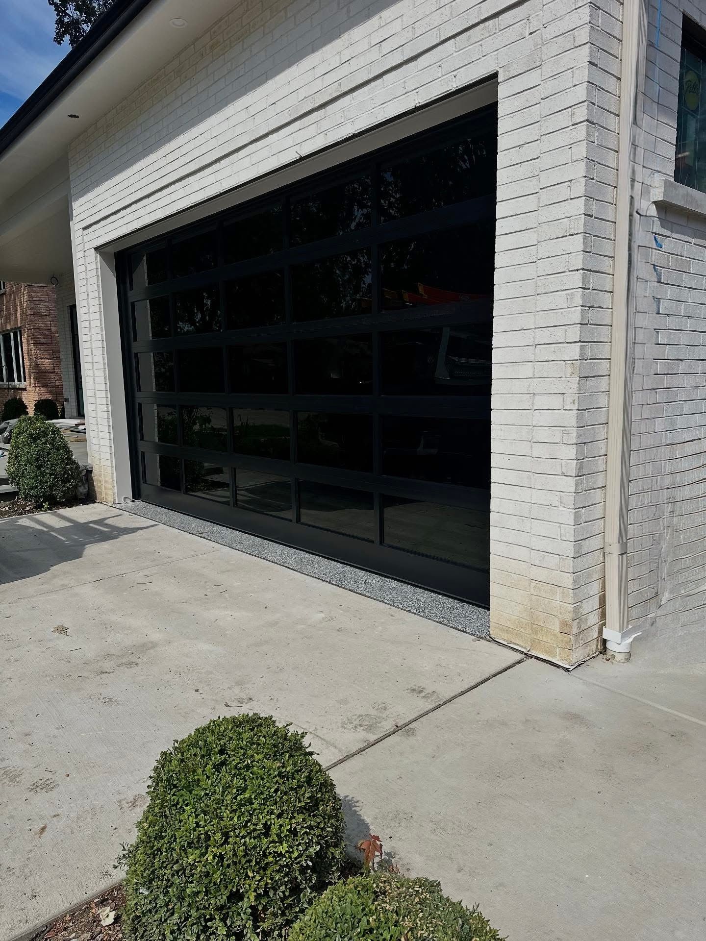Garage with black glass door and white brick exterior. Concrete driveway and small bushes.
