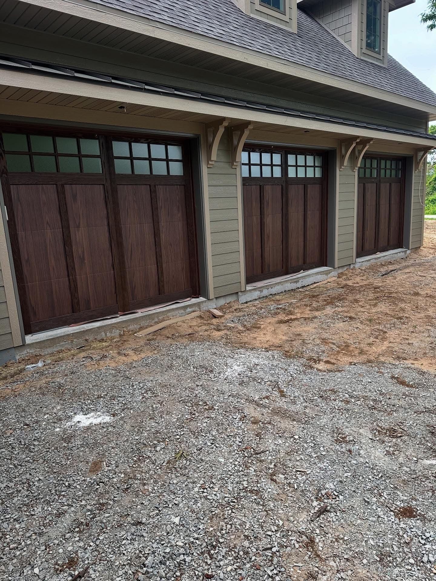 Three brown garage doors with glass panels under a beige house. Gravel foreground.