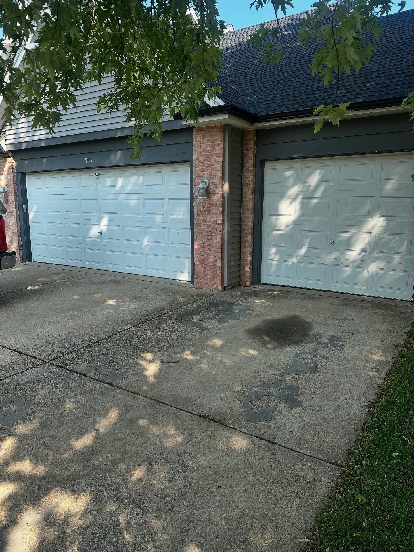 Two white garage doors with a brick column between them, on a concrete driveway, under a tree.