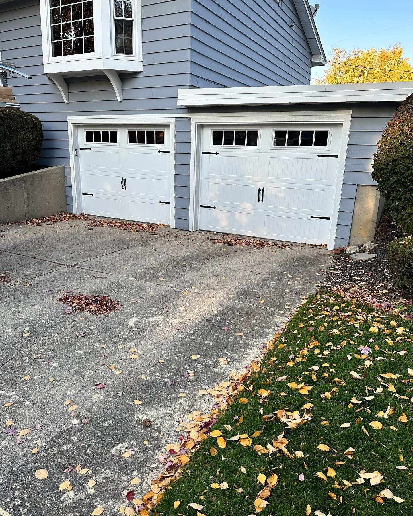 Two-car garage with white doors, on a driveway covered in leaves; part of a blue house.