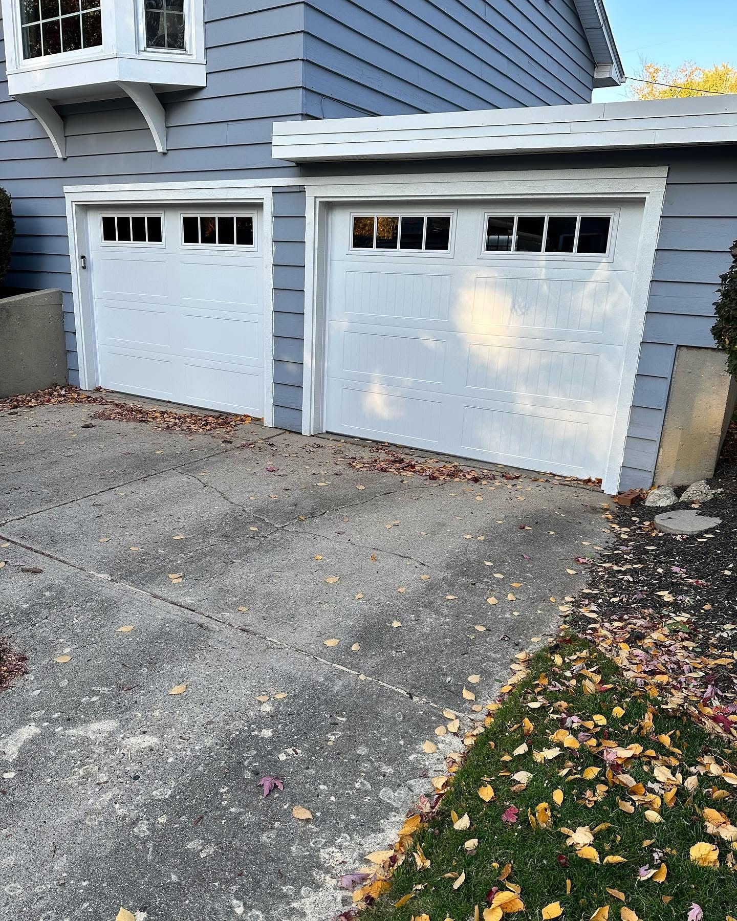 Two white garage doors on a blue house with a concrete driveway covered in leaves.