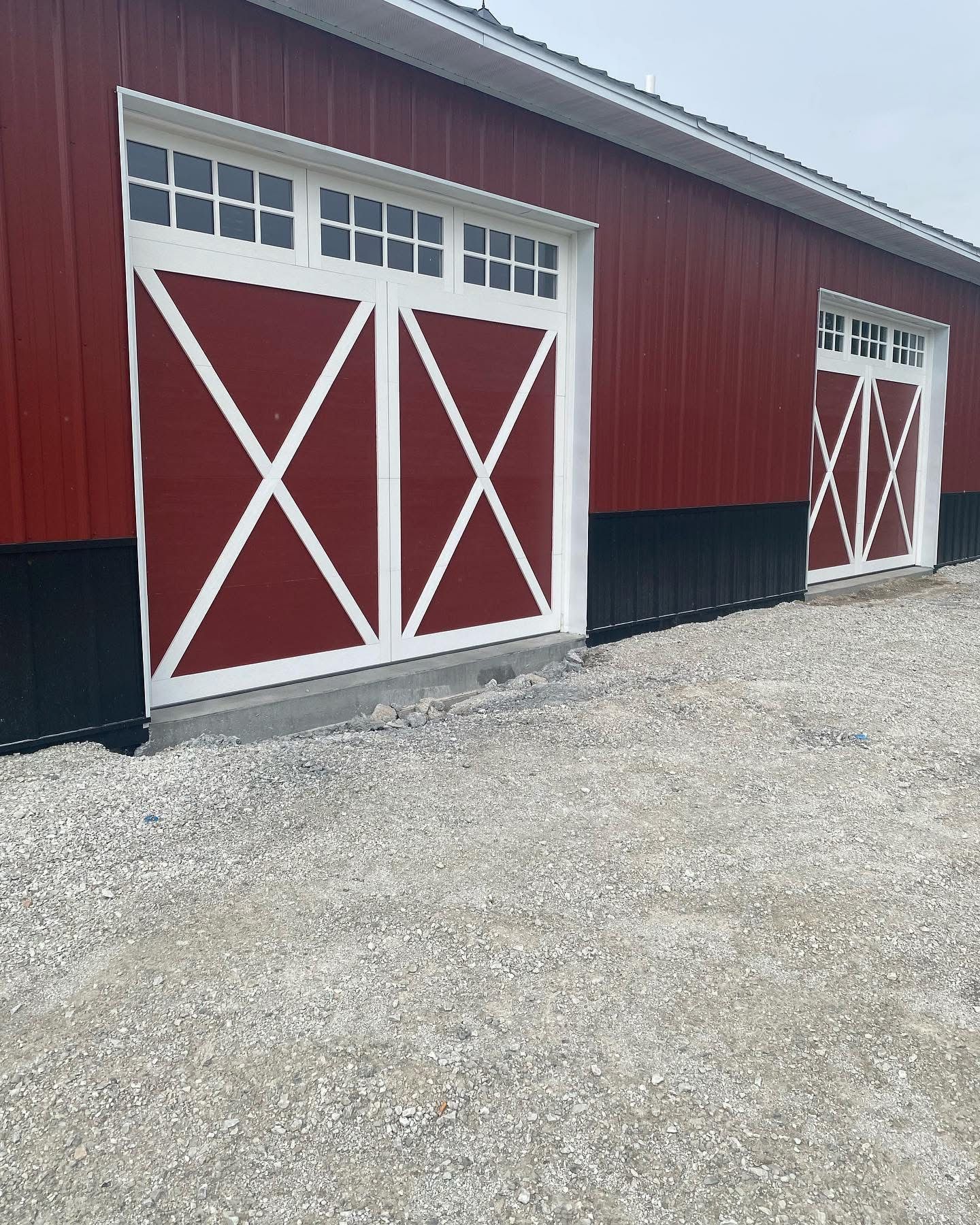 Red barn with white-trimmed doors featuring an 'X' design. Black base, white windows, gravel driveway.