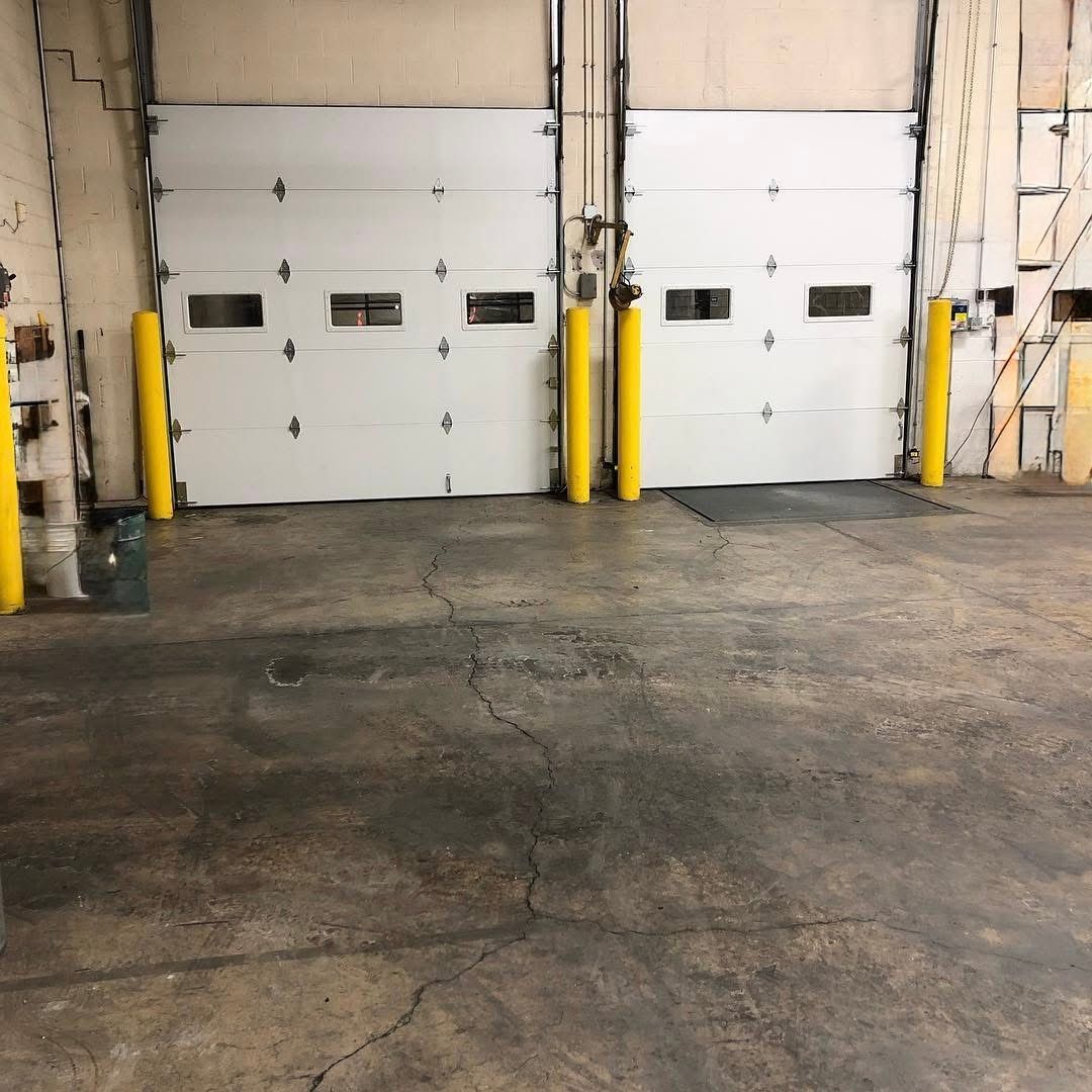 Two white garage doors in a warehouse, with yellow safety bollards on a cracked concrete floor.