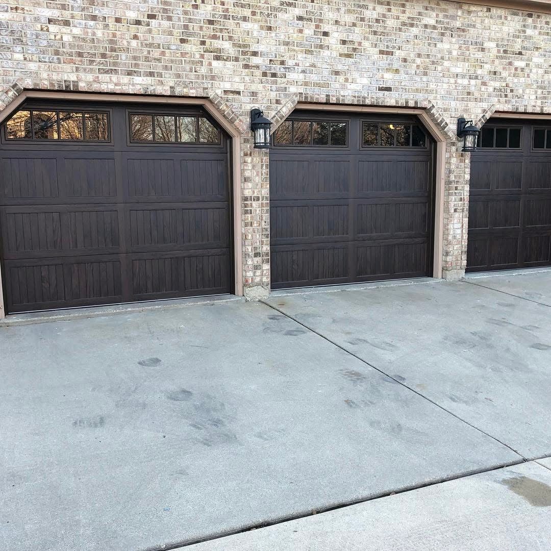Three dark brown garage doors with decorative windows on a brick house with concrete driveway.