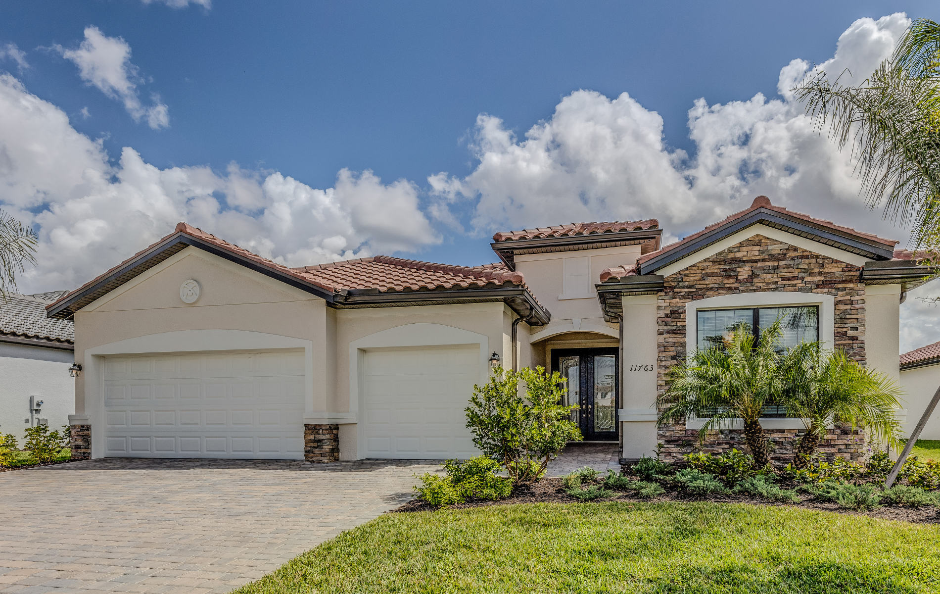 Beige stucco house with a tiled roof, two-car garage, and stone accent on a sunny day.