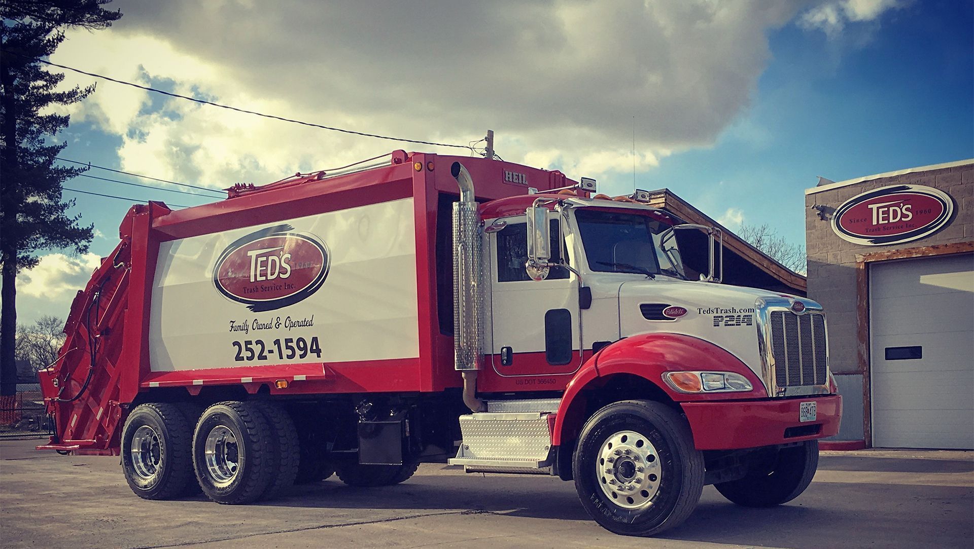 Red and white garbage truck parked beside a garage under a cloudy sky.