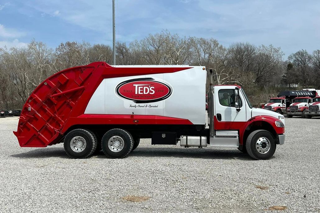 Red and white garbage truck parked on gravel lot with trees in the background.