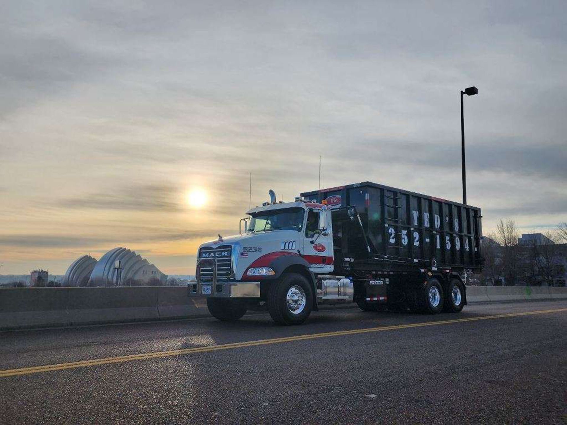 White dump truck parked on a road at sunset with a bridge in the background.