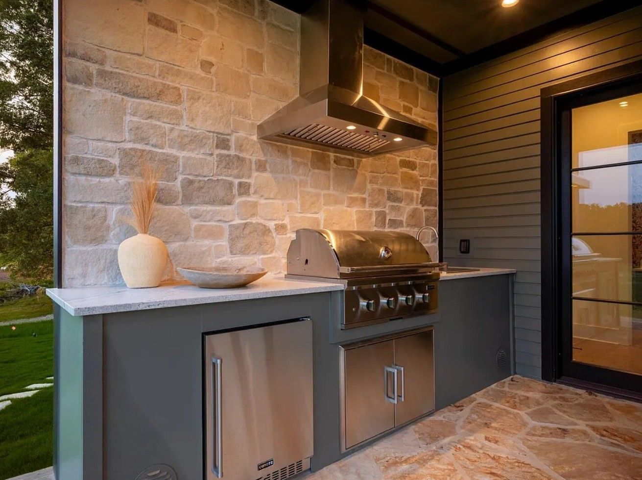 Outdoor kitchen with stone wall, stainless steel grill, fridge, and range hood.