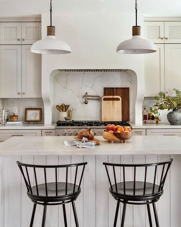 A kitchen with white cabinets , black chairs , a stove and a bowl of fruit on the counter.