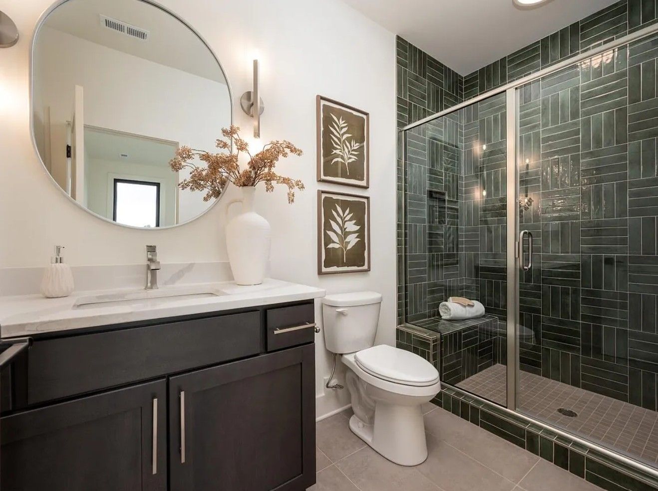Bathroom with dark vanity, green tiled shower, white toilet, round mirror, and botanical art.