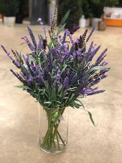 Lavender bouquet in a clear glass vase, purple flowers and green stems, on a beige floor.