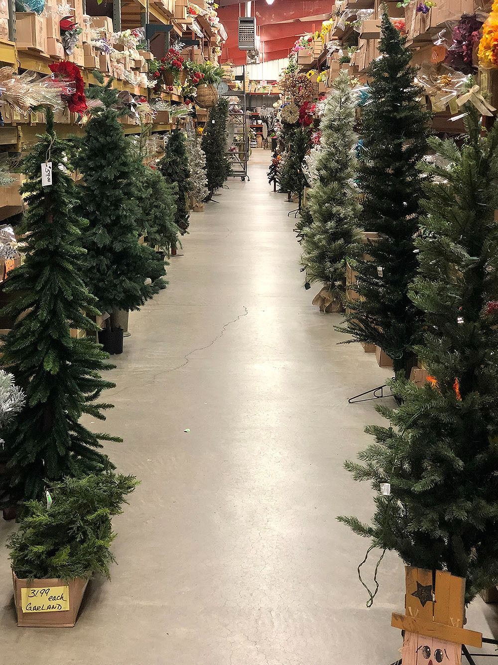 Rows of artificial Christmas trees in a store aisle; various sizes, green, with other holiday decorations in the background.