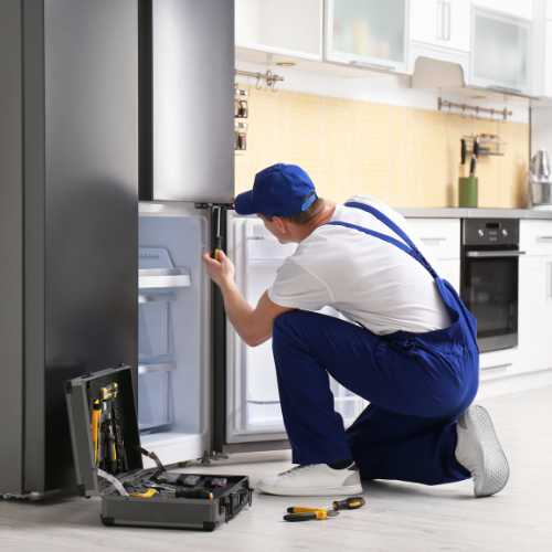 Appliance repair technician kneeling by an open refrigerator with tools in a bright kitchen.