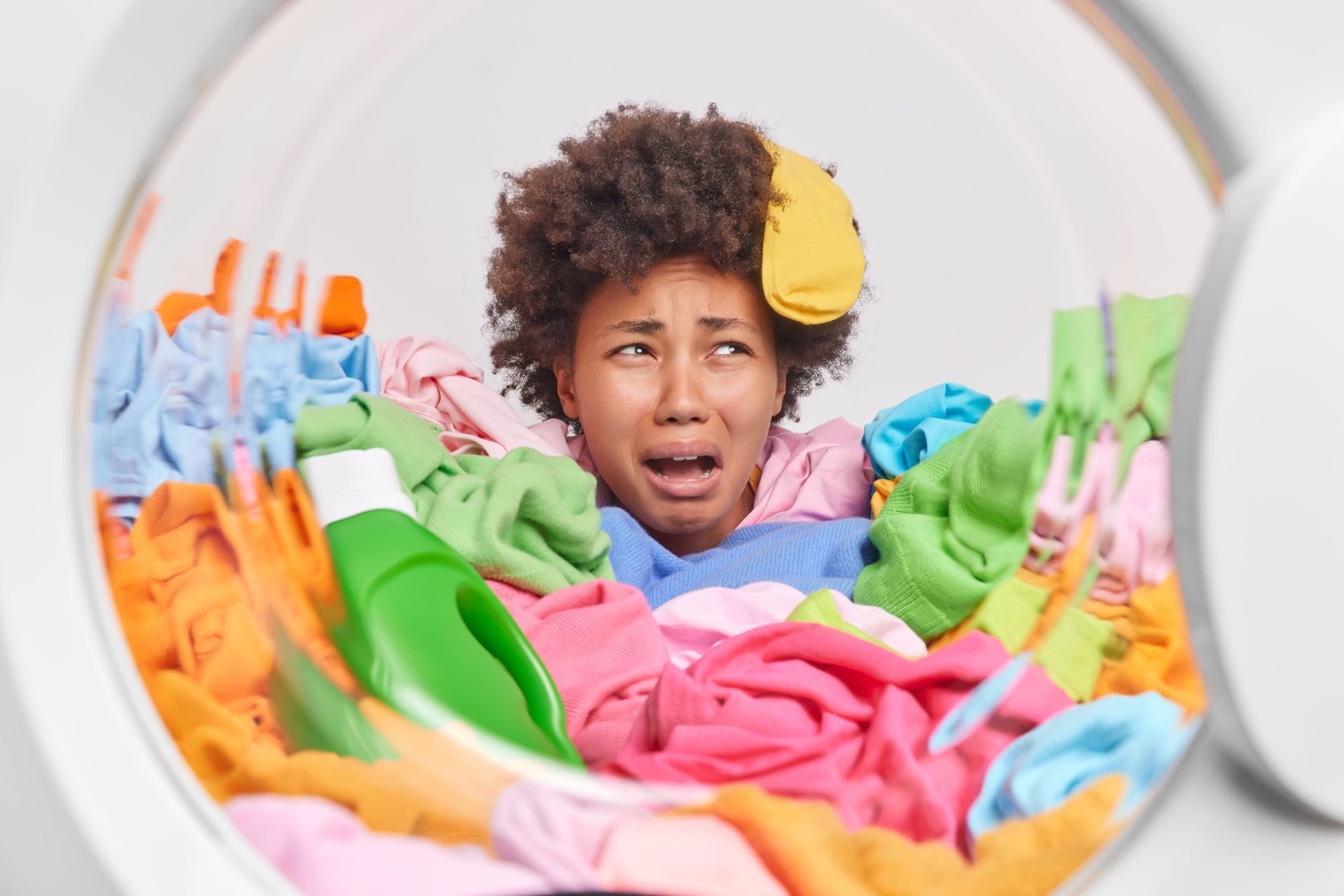 Person buried in a pile of colorful laundry inside a washing machine, looking frustrated