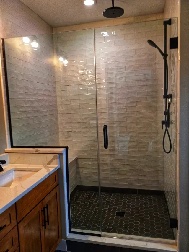 Modern shower with glass door and black fixtures; cream-colored textured tile walls, black hexagon tile floor; wooden vanity.