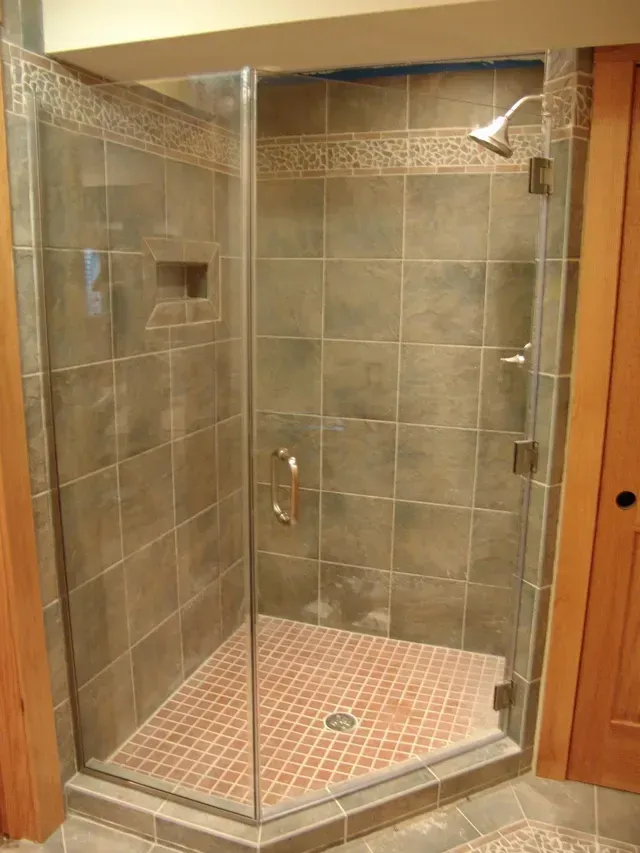Corner shower with glass doors, brown tile walls, and a patterned reddish-brown floor.