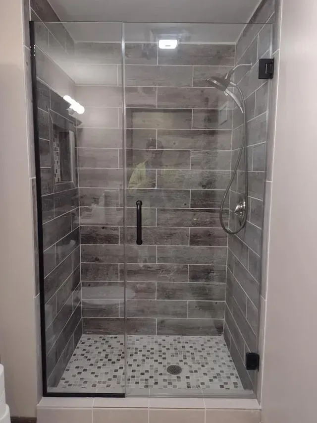 Shower stall with glass doors, gray tiled walls, pebble floor, and black hardware.