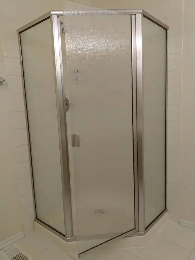 A corner shower enclosure with frosted glass panels and a silver frame, in a white tiled bathroom.