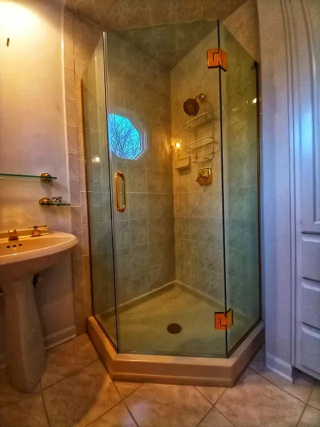Corner shower with glass doors and gold fixtures in a tiled bathroom, with a pedestal sink and cabinetry.