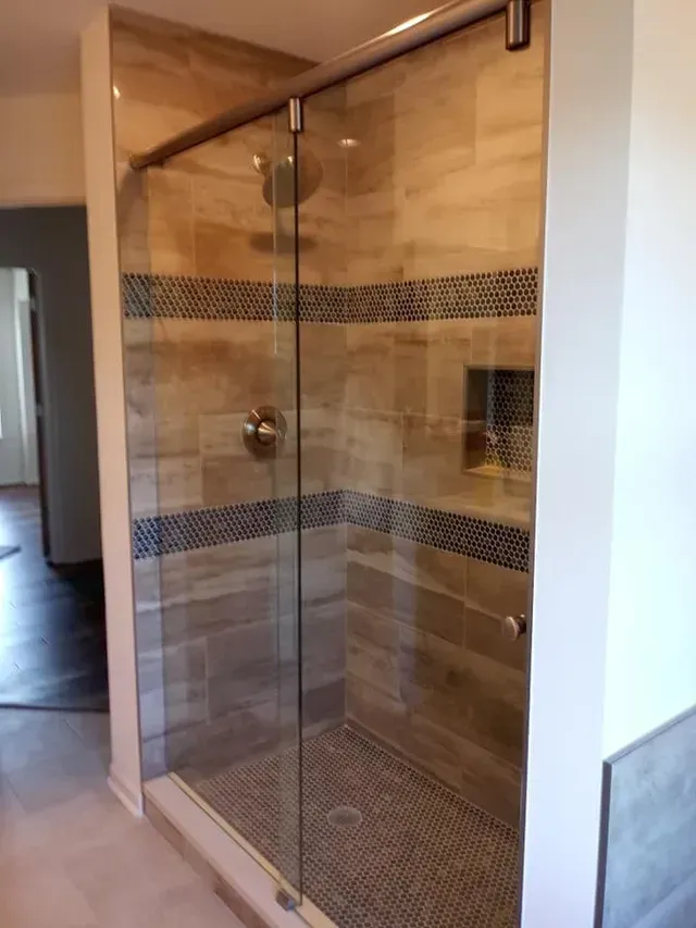 Glass-enclosed shower with beige and blue tiled walls. A stainless steel showerhead and door handle are visible.