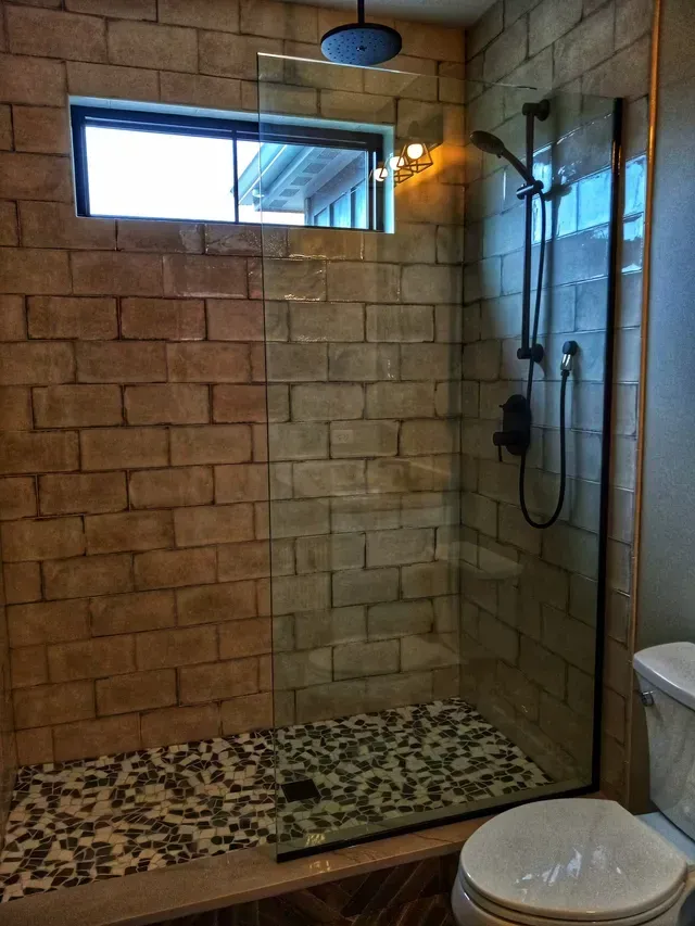 Shower with brick-like tile walls, pebble floor, glass door, window, and black fixtures.