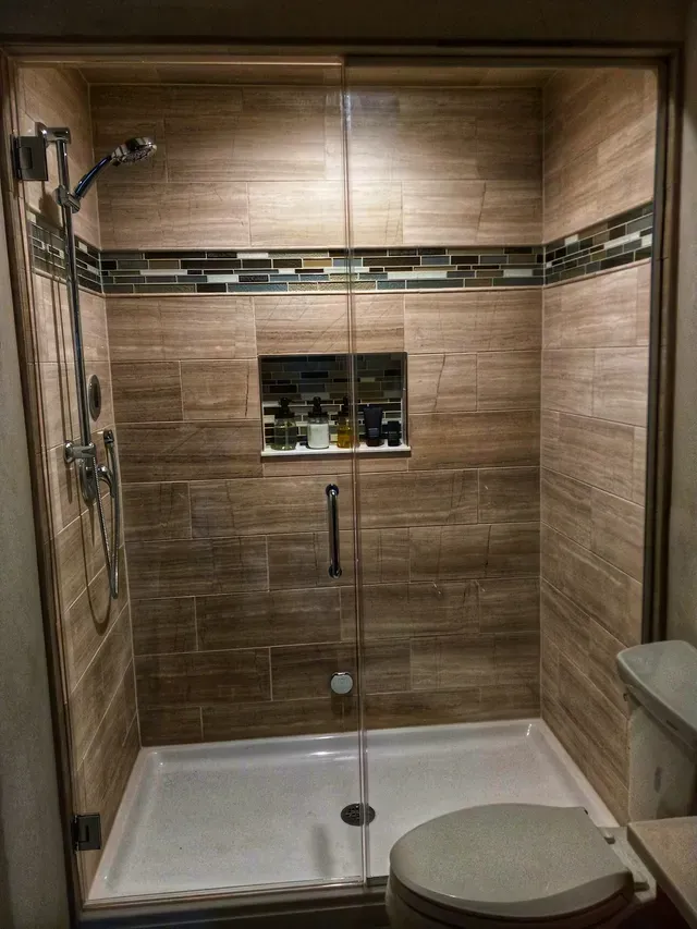 Shower with brown tile walls, glass door, and white shower pan. A small recessed shelf holds toiletries.