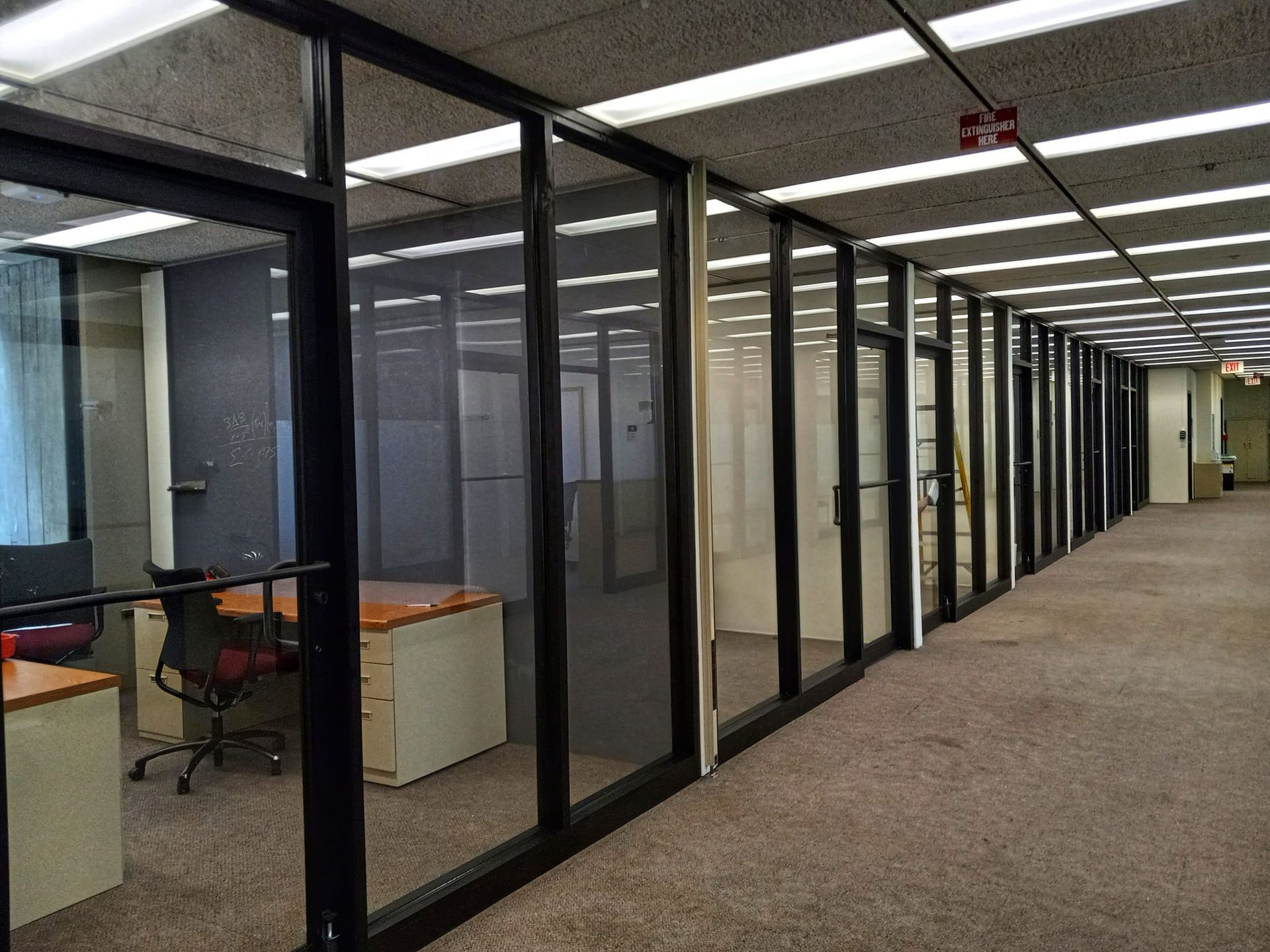 Office hallway with glass-walled rooms and fluorescent lighting. Beige carpet, black frames, and office furniture visible within.