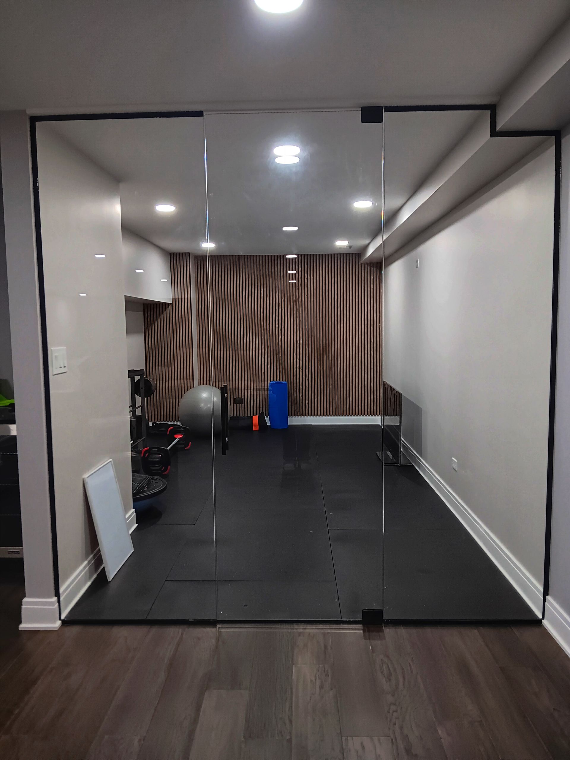 Glass-walled home gym with black mats, exercise equipment, and wood-paneled wall backdrop.