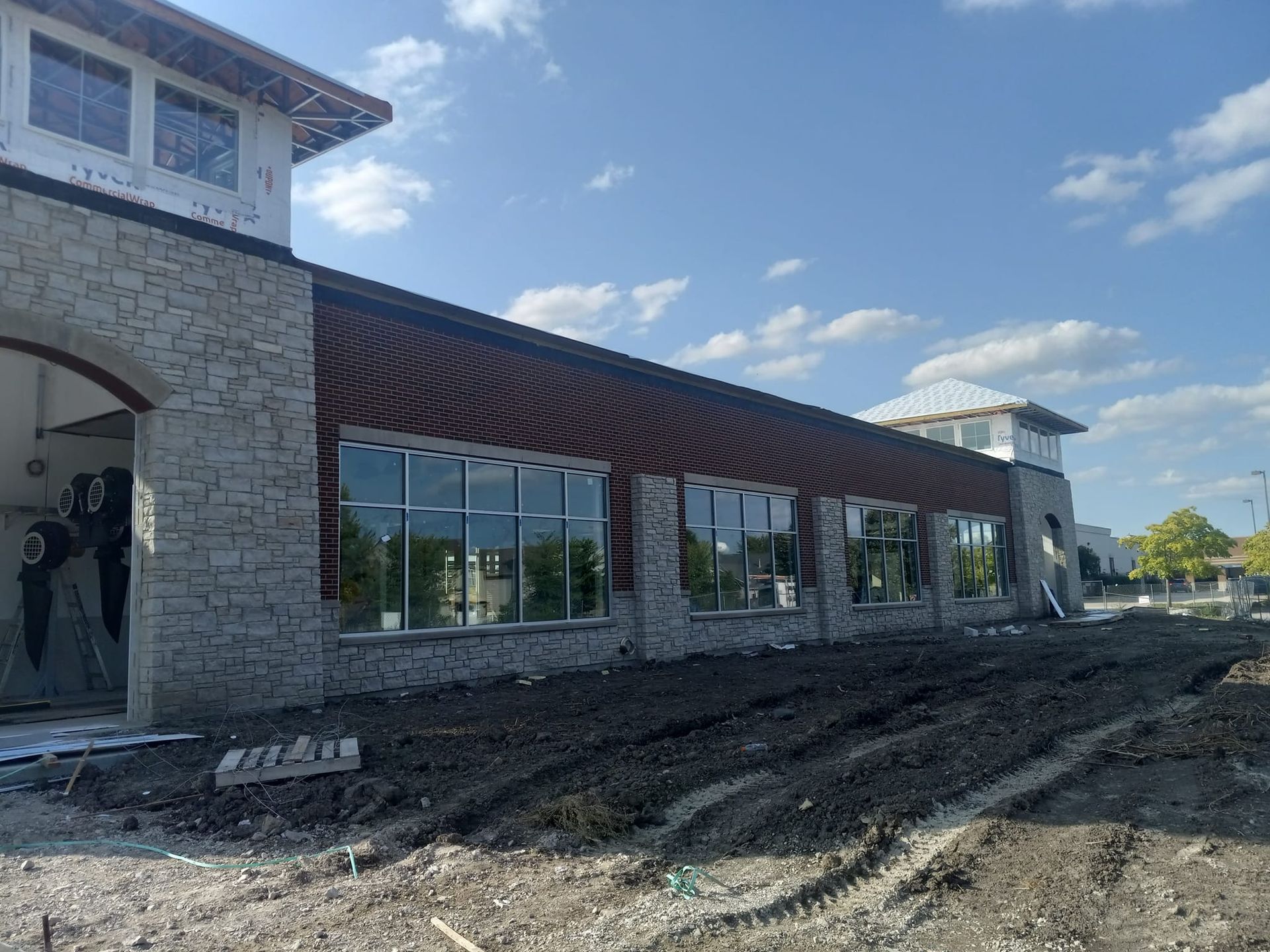 Building exterior under construction with stone facade, large windows, and dirt ground.