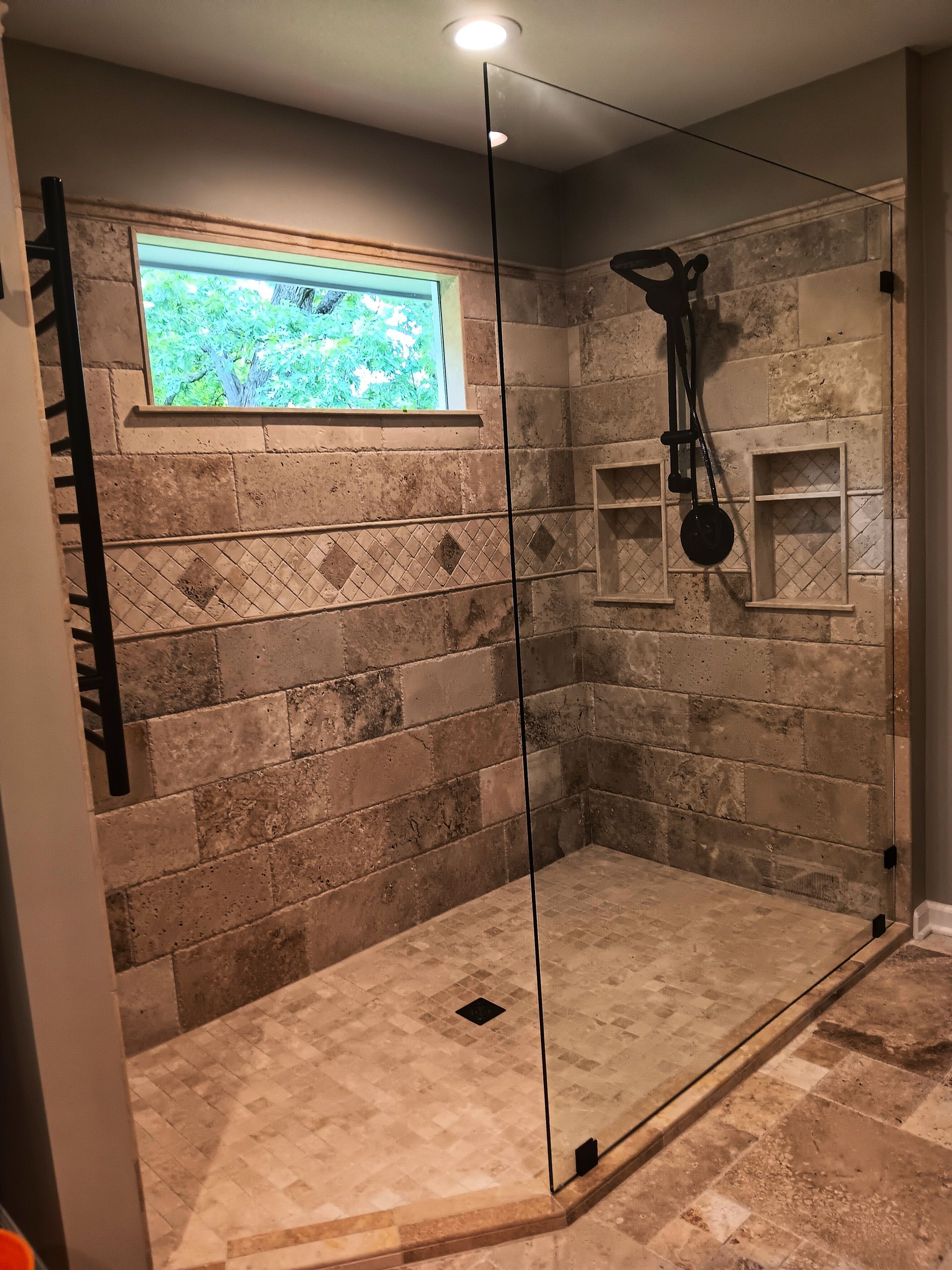Shower stall with tan stone tiles, glass walls, a rain showerhead, and a small window.