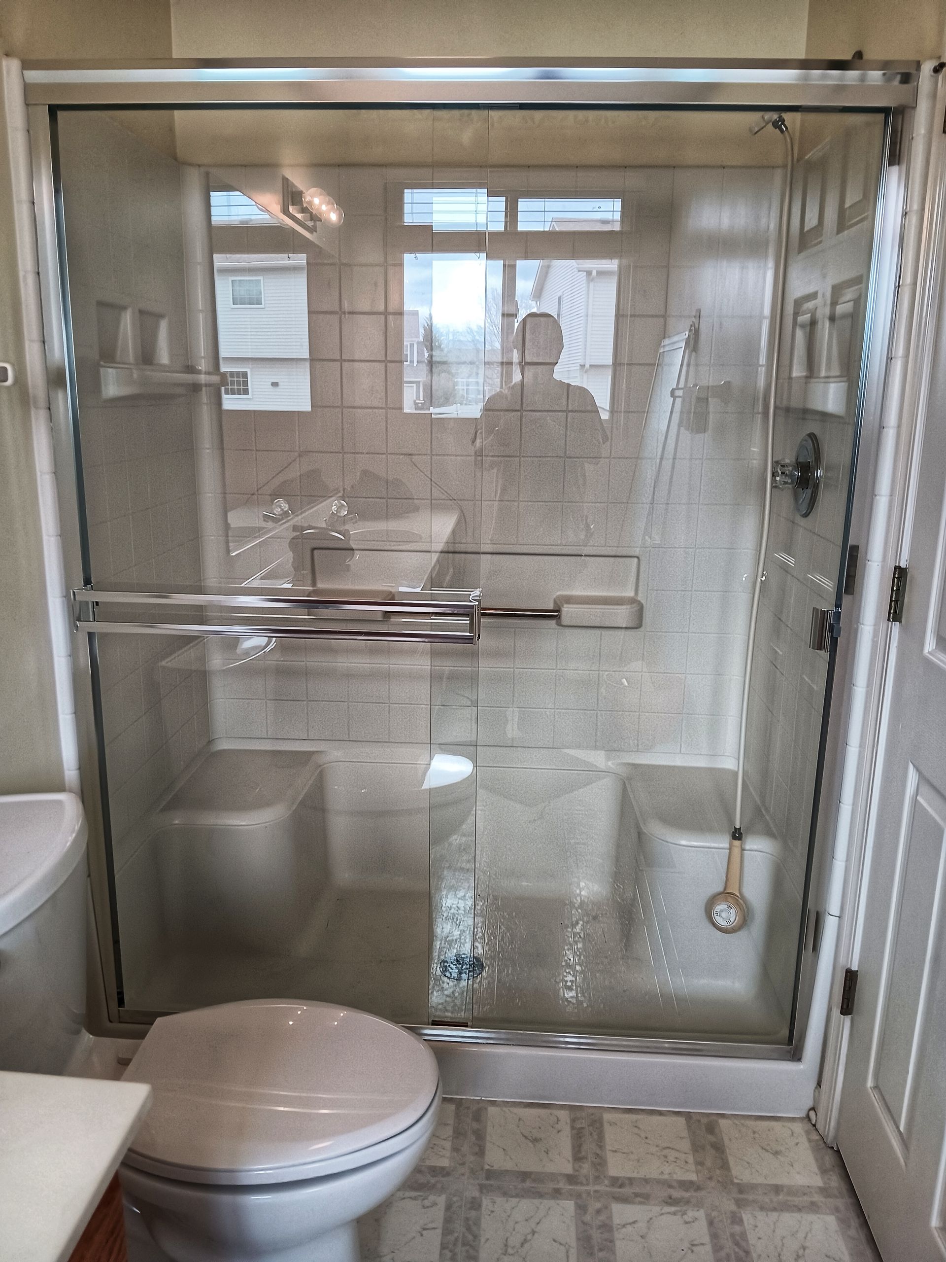 A clear glass shower with water droplets, next to a white toilet in a bathroom with beige tiles.