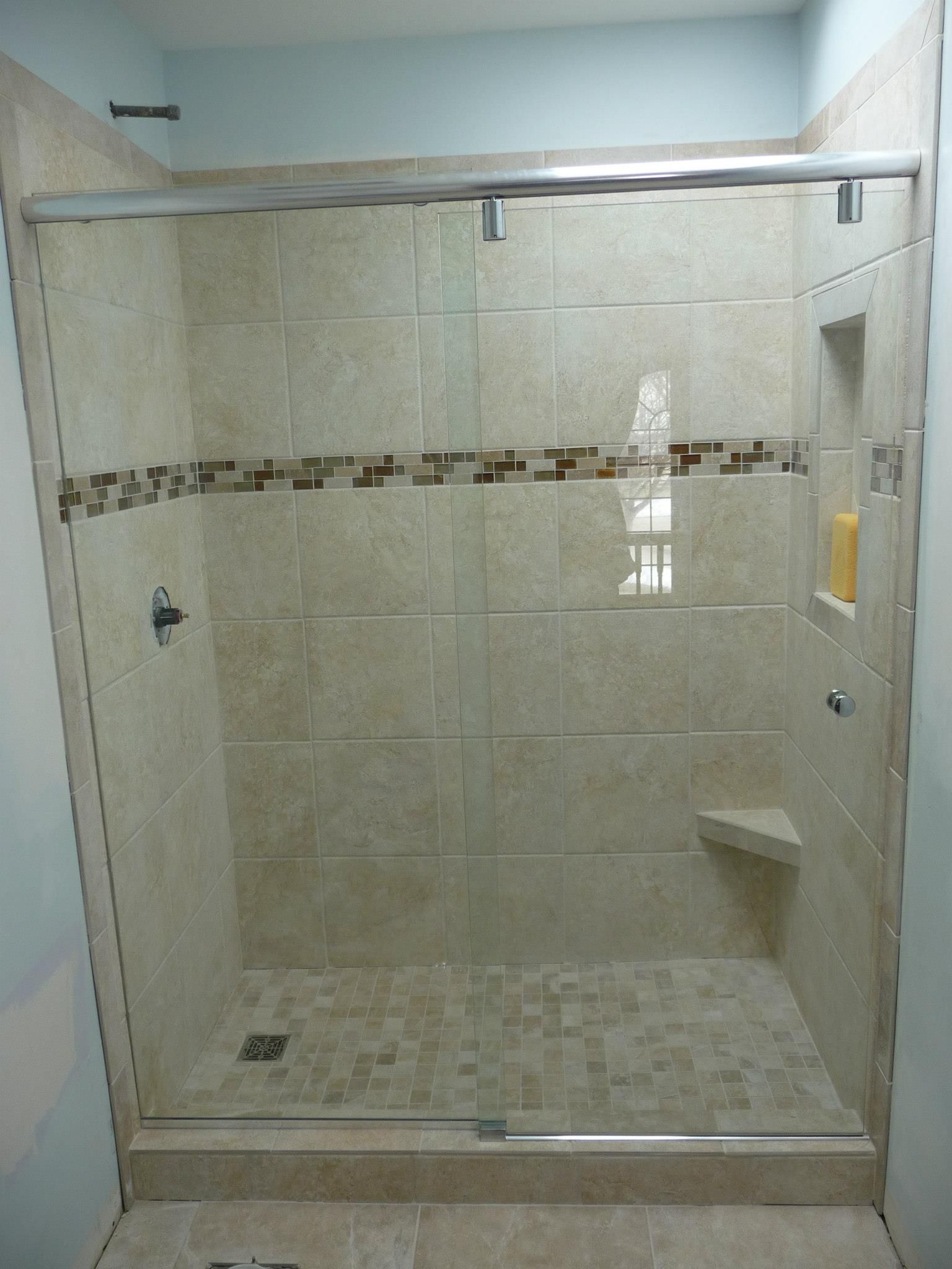 Tiled shower with glass doors. Beige tiles, brown accent stripe. Built-in shelf, corner seat. Chrome fixtures.