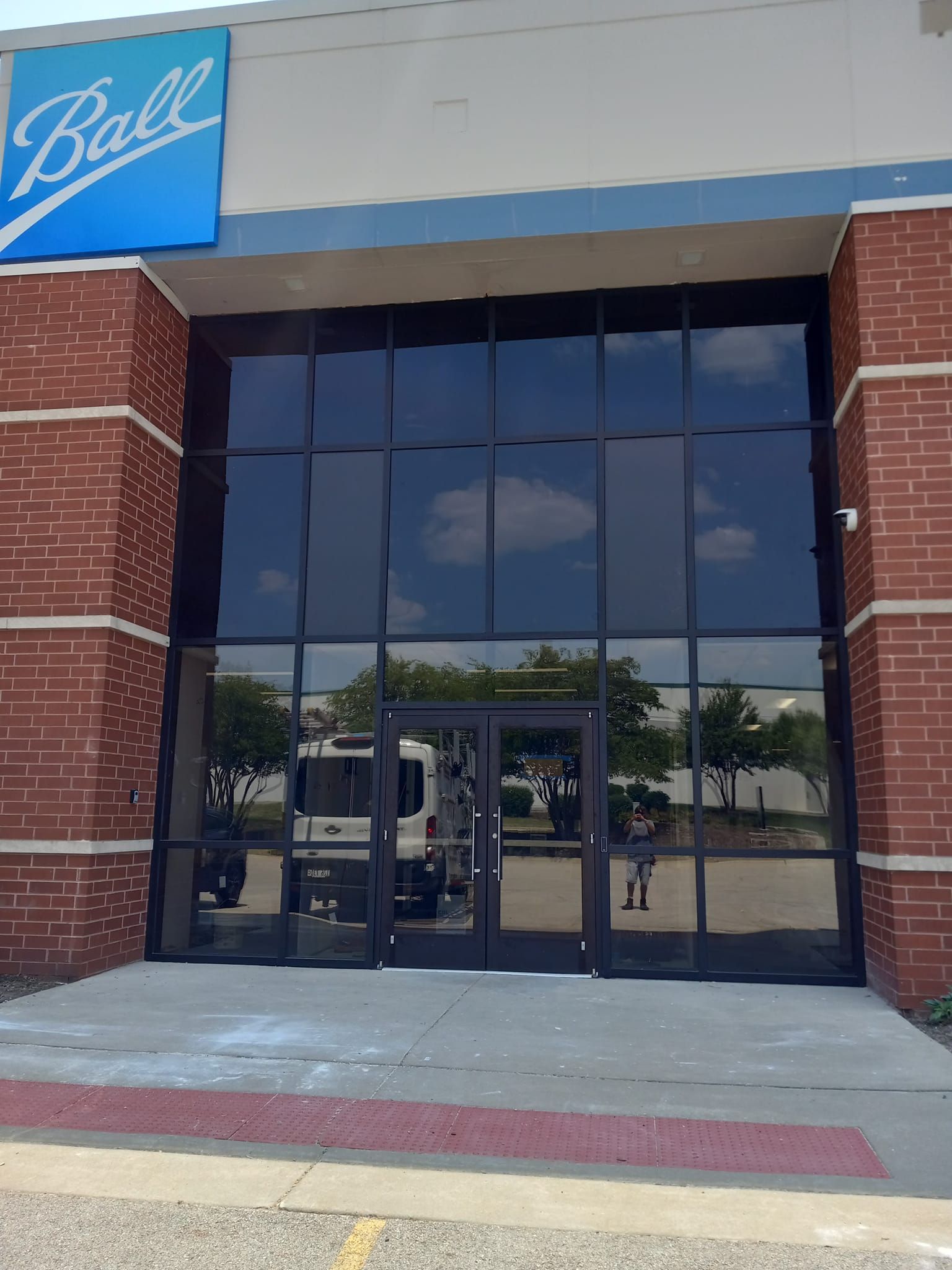 Ball Corp. building entrance with glass doors and windows, blue logo, brick exterior, and a reflecting sky.